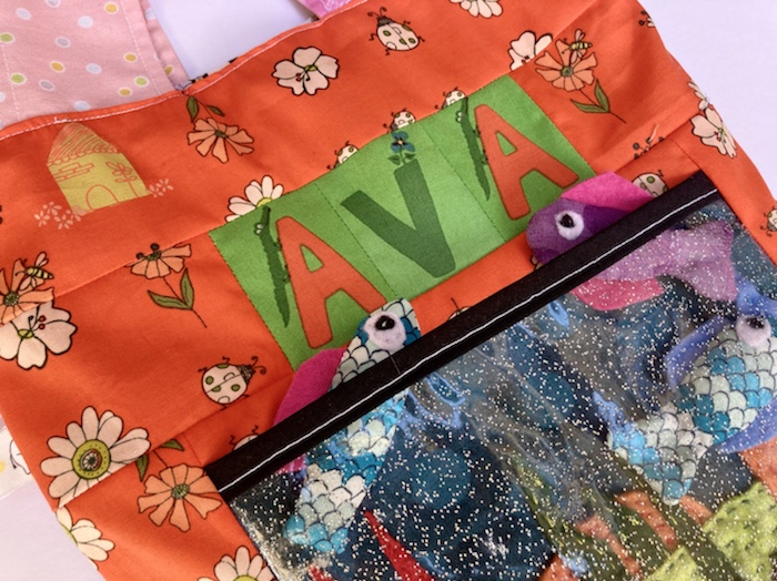 Close up of the handmade tote bag - it says AVA and has a fish tank on the front with Tiddly Fish poking out the pocket